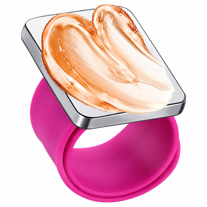 A square face mask with a silver metallic border and a glossy surface displaying a swirled smear of warm beige and peach-toned cream makeup, mounted on a wide, flexible magenta pink silicone ring band, shown against a plain white background.